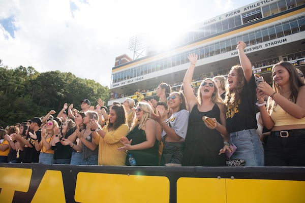 This example image is being used to help people understand how to write alternative text. The image shows students dressed in black and gold with App State logos present on their clothing. They are cheering and clapping in the stands (student section) of Kidd Brewer Stadium. The students are in the forefront, and the background of the image shows the top of the stadium with private boxes and "Mark E. Ricks" showing.